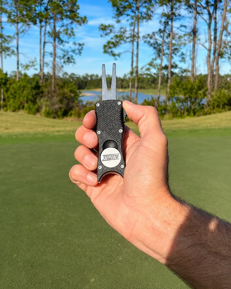 A hand holding the Stiletto Black Nylon switchblade ball mark repair tool with its textured black handle and forked tip extended, overlooking a golf course