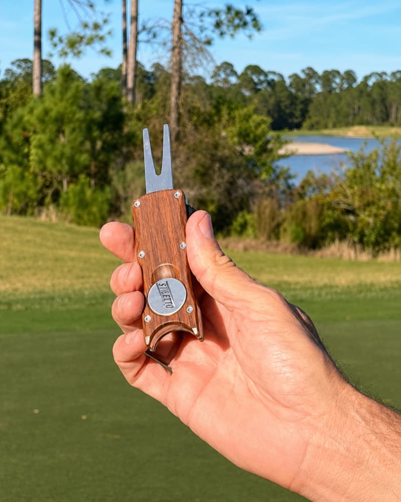 A hand holding the Stiletto Prestige switchblade ball mark repair tool with its wood handle and forked tip extended, overlooking a golf course